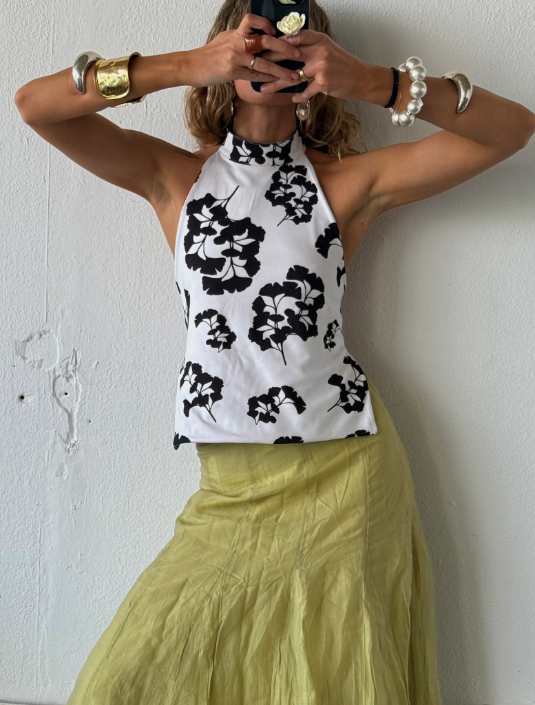 A woman in a floral top and skirt smiles as she takes a selfie outdoors