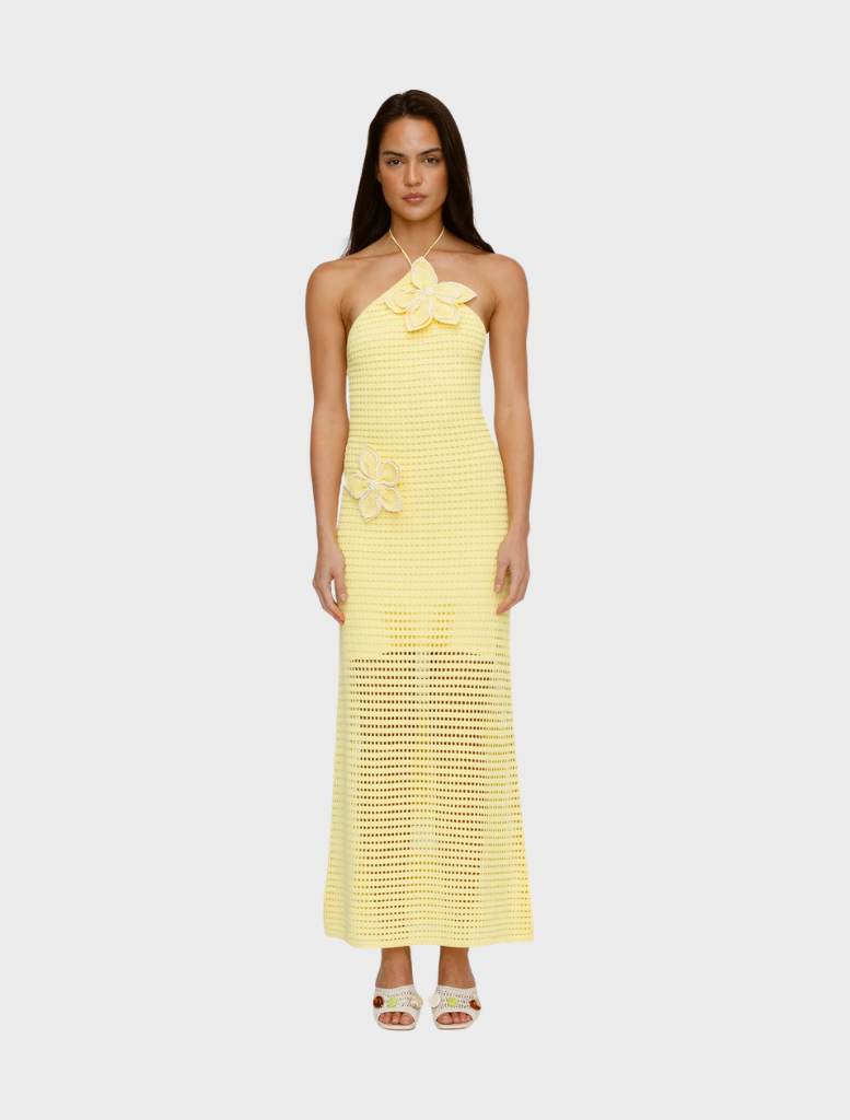 Woman in a yellow crochet halter dress with floral appliqués, standing against a plain background. The look is summery and elegant, with a relaxed vibe.