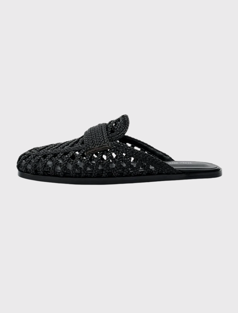 Krissy is a black woven flat mule featuring a wide crossover strap, closed toe, and open back design for easy slip-on wear.