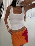 A woman wearing a white tank top and an orange skirt stands confidently, showcasing her vibrant outfit.
