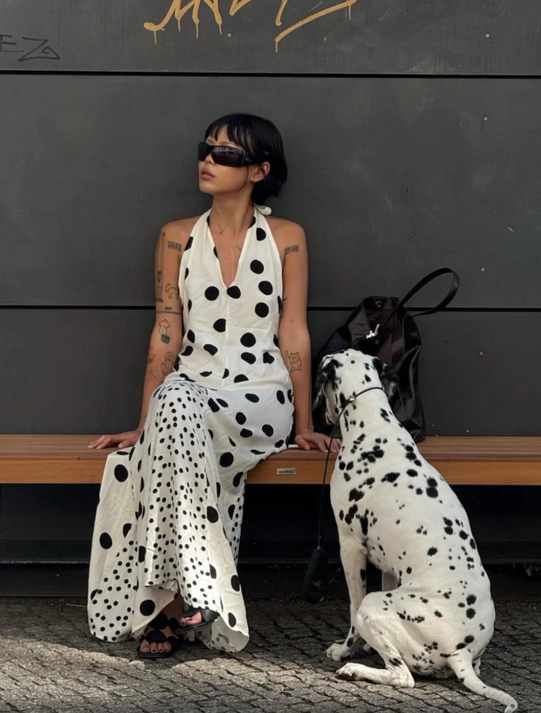 A woman in a polka dot dress stands in front of a mirror, adjusting her hair and admiring her reflection.