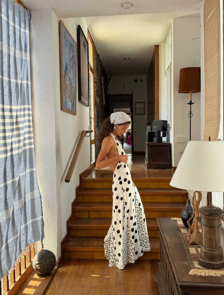 A woman in a polka dot dress stands in front of a mirror, adjusting her hair and admiring her reflection.