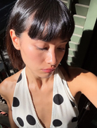 A woman wearing a polka dot dress stands before a mirror, looking at her reflection with a smile.