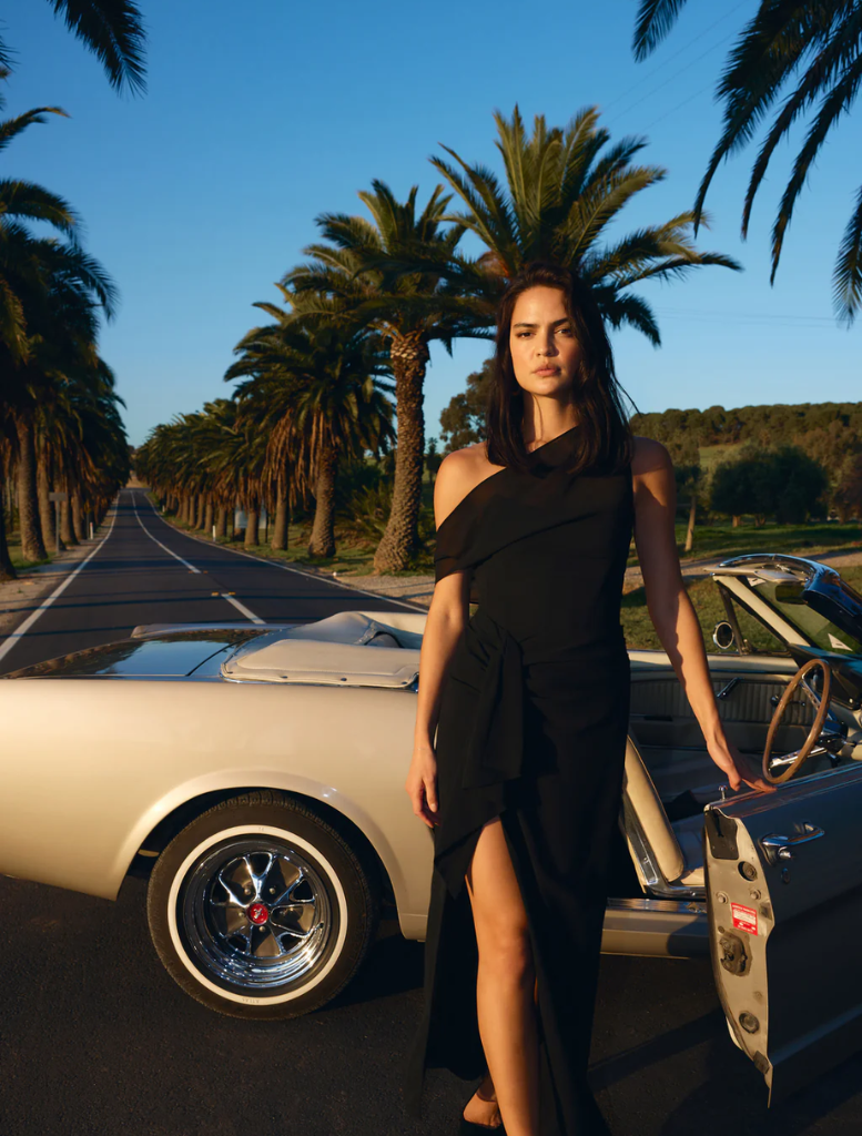 A woman in a black dress stands beside a classic car, showcasing elegance and vintage style.