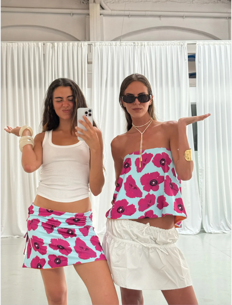 Two women wearing matching floral skirts and white tops stand together, smiling at the camera.