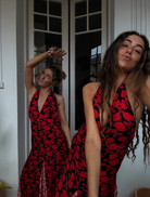 A woman in a striking red and black dress stands atop a stone wall, blending elegance with a rugged landscape.