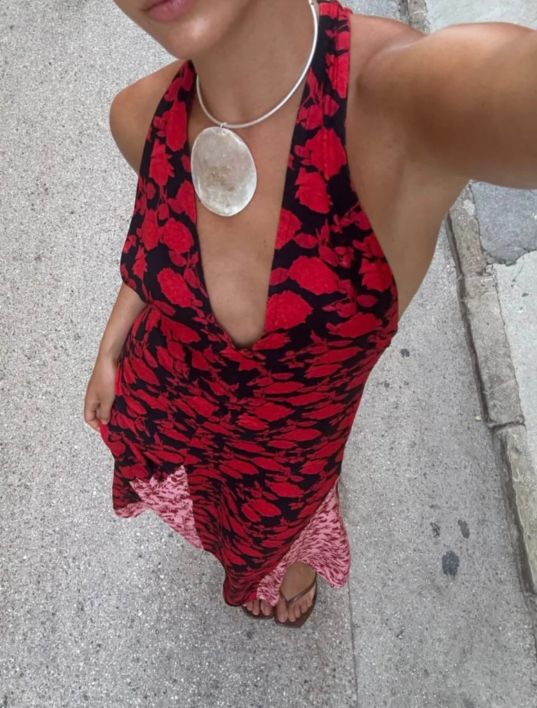 A woman in a striking red and black dress stands atop a stone wall, blending elegance with a rugged natural environment.