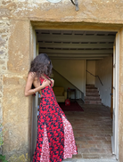 A woman in a red and black dress stands on a stone wall, exuding confidence in a picturesque outdoor environment.