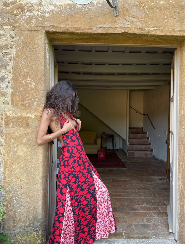 A woman in a red and black dress stands on a stone wall, exuding confidence in a picturesque outdoor environment.