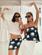 Two women wear white tank tops and skirts adorned with white flower, standing together in a bright outdoor setting.
