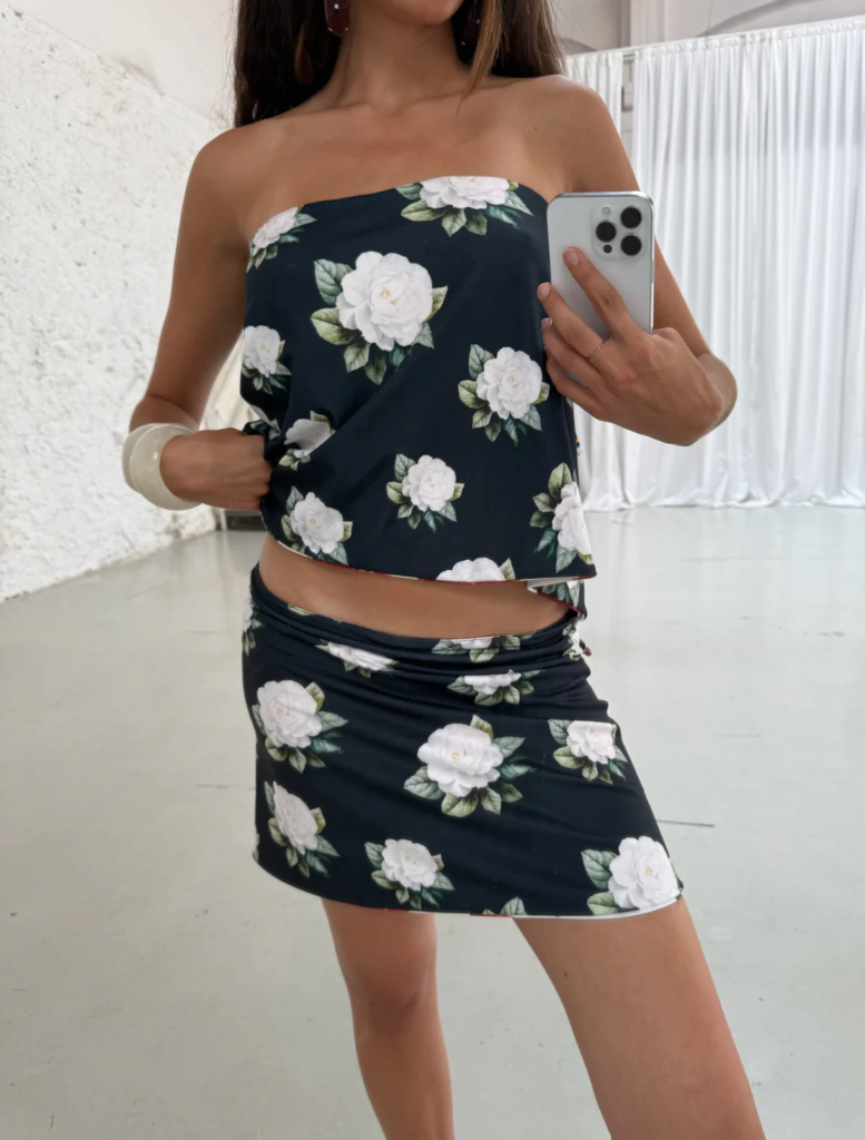 A woman in a floral dress smiles as she takes a selfie outdoors, surrounded by greenery.