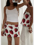 Two women wear white tank tops and skirts adorned with strawberries, standing together in a bright outdoor setting.