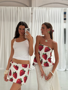 Two women in matching white tank tops and skirts featuring strawberries pose together, showcasing a cheerful summer vibe.