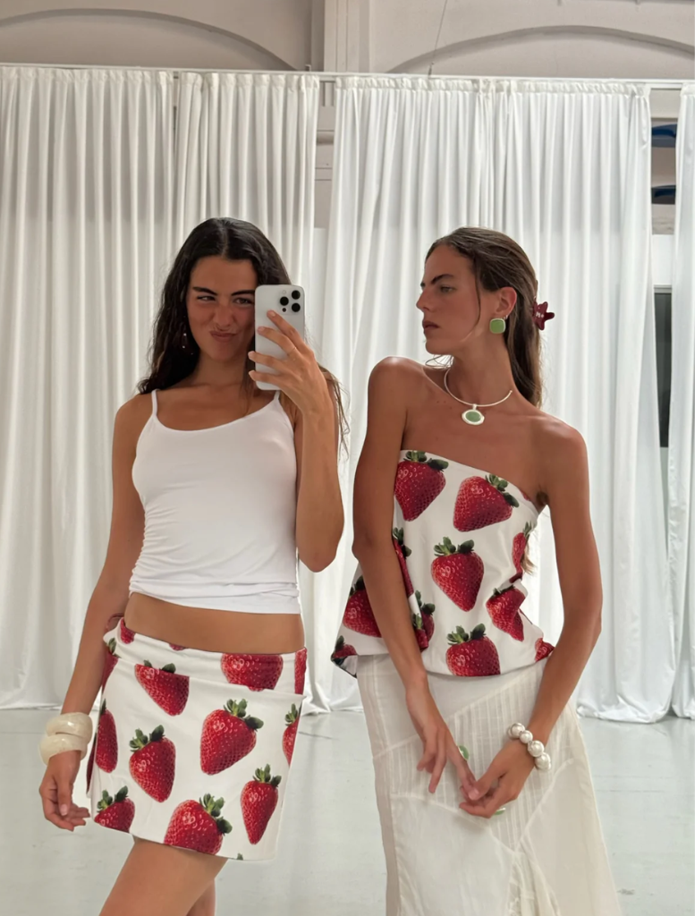 Two women in matching white tank tops and skirts featuring strawberries pose together, showcasing a cheerful summer vibe.