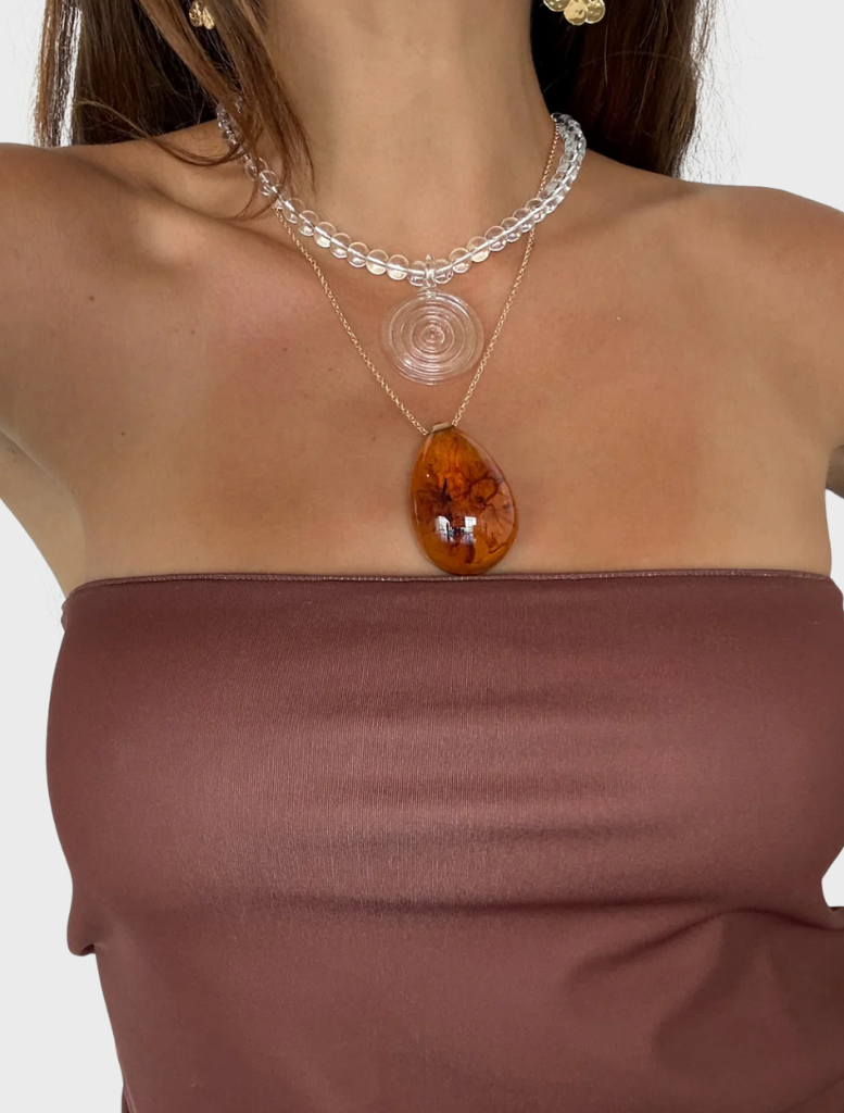 A woman wearing a polka dot dress and the Elara Necklace in clear sits gracefully on a window sill.