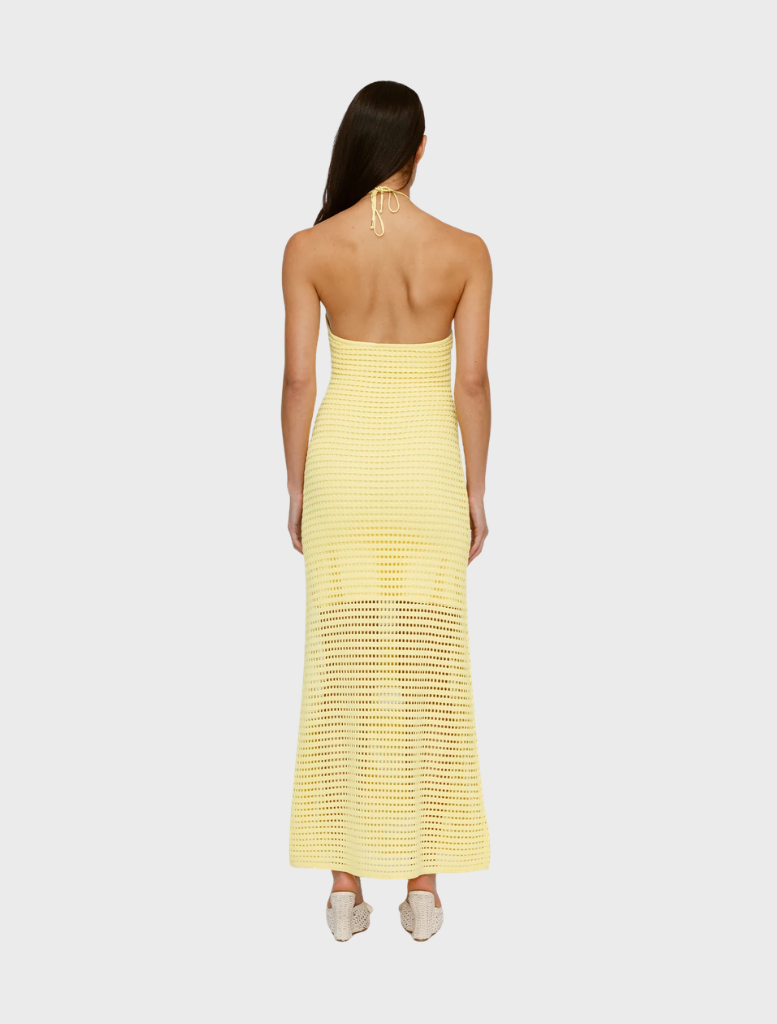 A woman with long hair is seen from behind wearing a light yellow, backless crochet halter dress and white slip-on shoes. The tone is relaxed and summery.