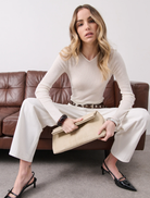 Woman in a white outfit sitting on a brown leather couch holding a beige clutch.