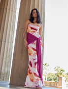 A woman in a pink mineral print dress stands gracefully in front of tall pillars, showcasing elegance and style.