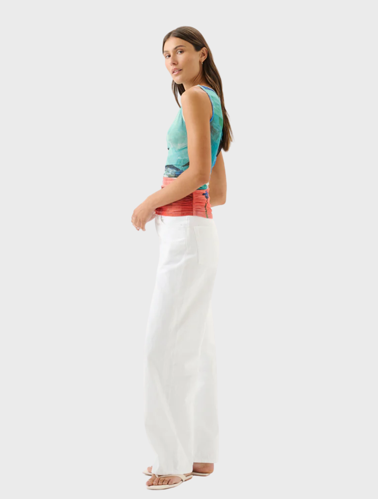 A woman wearing white trousers and a colorful blouse poses, displaying her trendy and cheerful outfit.
