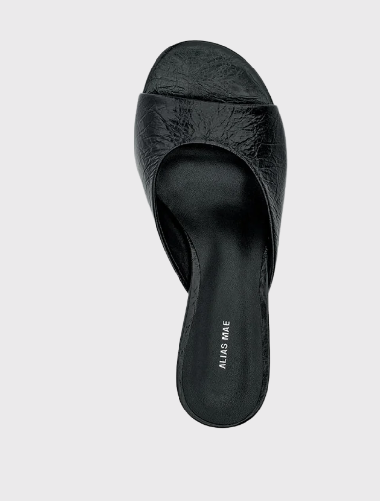 Black oily crinkle leather mule with a wide front strap, sculptural wedge heel, and open-back slip-on style.