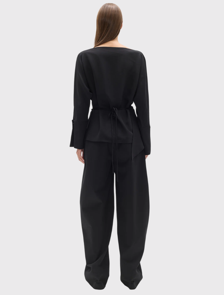 Novalyn Top in black with a boat neckline, draped front, self-tie waist, relaxed fit, long sleeves, and buttoned cuffs.
