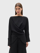 The Novalyn Top in black features a boat neckline, draped front, self-tie waist, relaxed fit, long sleeves, and buttoned cuffs.