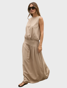 A woman in a tan maxi dress, the Delphine Skirt in Nude, poses gracefully, highlighting the dress's elegant silhouette.