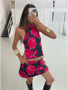 A woman in a floral dress smiles as she takes a selfie outdoors, surrounded by greenery.
