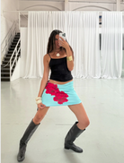 A woman wearing a skirt and boots smiles while taking a selfie outdoors.
