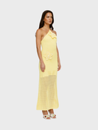 A woman in a sleeveless, halter-neck, yellow crochet dress adorned with floral details stands sideways. She has long hair and wears white open-toed heels.