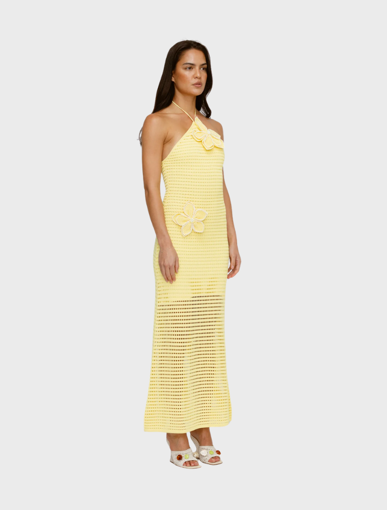 A woman in a sleeveless, halter-neck, yellow crochet dress adorned with floral details stands sideways. She has long hair and wears white open-toed heels.