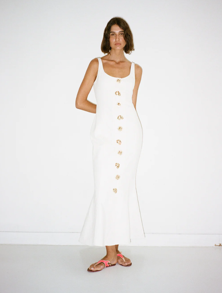 A woman wearing an ivory Sibel Denim dress and sandals stands elegantly, showcasing her stylish summer outfit.