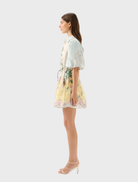 A model displays the Berkley Mini Dress - Utopia, adorned with a lively print of palm trees and floral designs.